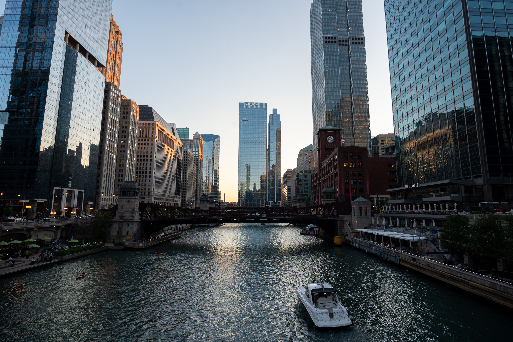 The Chicago River at dusk, flowing between downtown high-rise buildings with a bascule bridge crossing mid-frame and a small white boat cruising toward the camera.